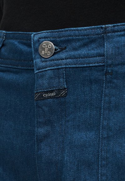 Gros plan sur la ceinture d'un jean en denim bleu foncé avec un bouton en métal et une étiquette noire, tous deux arborant le mot "CLOSED".