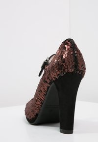 Chaussure à talon haut recouverte de sequins bruns avec un dos en suède noir, bout pointu et une bride ajustable à la cheville. Talon surélevé pour plus de hauteur.