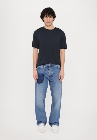 T-shirt noir à manches courtes, jeans bleus clairs amples avec une poche en patch foncé, et baskets blanches sur fond uni.