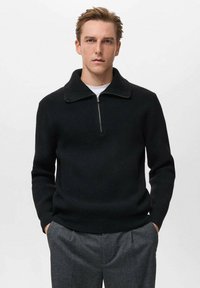 Pullover nero a coste con colletto alto e chiusura con zip. Il design presenta maniche lunghe e una silhouette aderente. Indossato con pantaloni grigi.