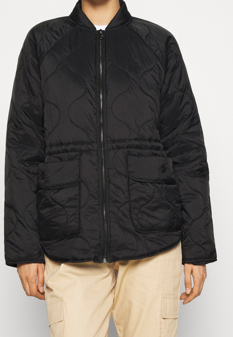 Svart vadderad jacka med dragkedja framtill, rund krage, två fickor med knappslå och en texturerad yta. Har en avslappnad passform och lättviktsmaterial.