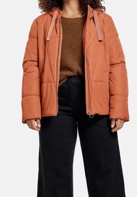 Žena v hnědé péřové bundě s rezavými odstíny, oblečená na hnědém pleteném svetru a černých vysokých kalhotách, stojí na bílém pozadí.