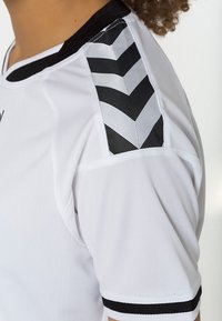 Acercamiento a la manga de una camiseta deportiva blanca con franjas en zigzag en blanco y negro en el hombro y ribetes negros en el cuello y el puño.