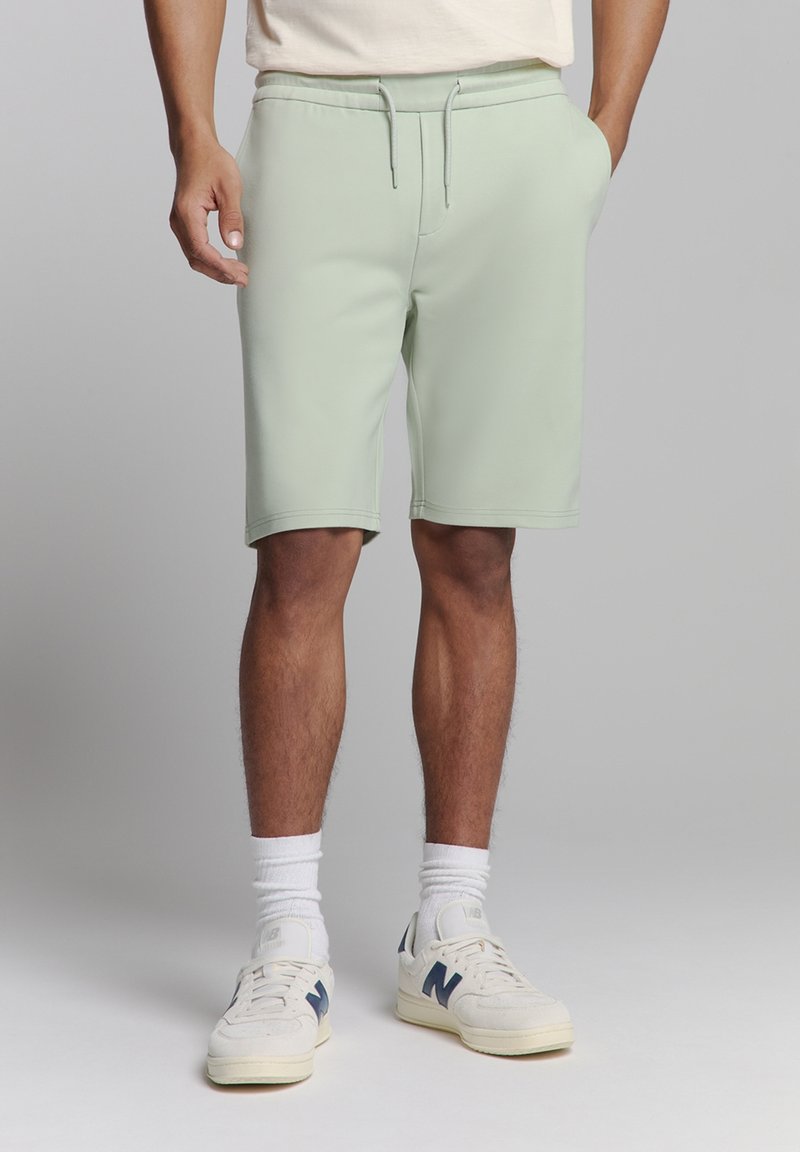 Licht mintgroene shorts met een elastische tailleband en trekkoord, met een gladde textuur en een knielange pasvorm. Gemaakt in combinatie met witte sokken en sneakers.