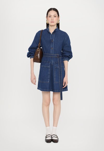 Rochie din denim cu guler, mâneci lungi, două buzunare frontale, talie marcată cu brâu, de culoare albastru închis. Purată cu șosete albe și pantofi negri.