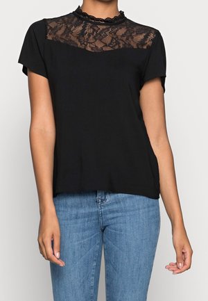 Blouse noire à manches courtes avec empiècement en dentelle et col montant, associée à un jean bleu portée par une personne debout devant un fond clair uni.