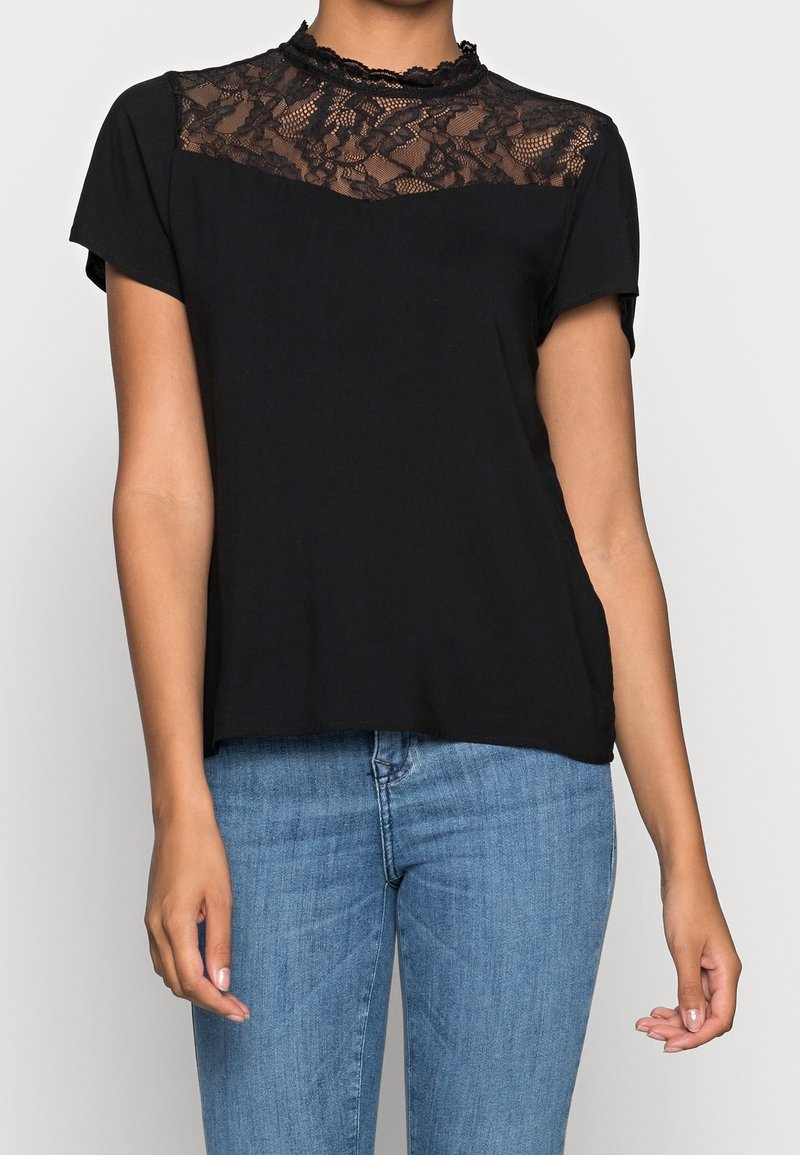 Blouse noire à manches courtes avec empiècement en dentelle et col montant, associée à un jean bleu portée par une personne debout devant un fond clair uni.