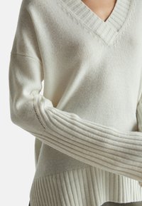 Weißer Strickpullover mit V-Ausschnitt, mit gerippten Ärmeln und strukturiertem Körper. Weicher, leichter Stoff mit dezenten vertikalen Musterakzenten.