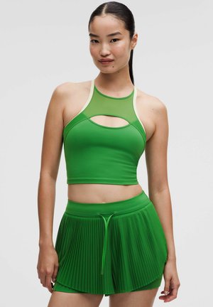 Femme aux cheveux foncés portant un crop top vert avec découpe et un short plissé vert assorti sur un fond gris uni.