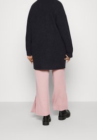 Længere marineblå sweater med ribbet tekstur, parret med lyserøde vidde bukser med et subtilt mønster og sideslids, afsluttet med sorte støvler.