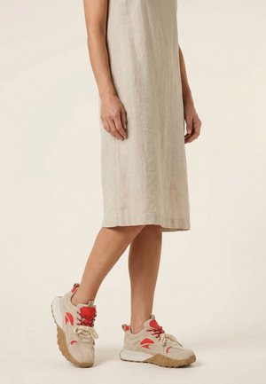 Onderlichaam van een persoon die een beige jurk tot op de knie en beige sneakers met rode accenten draagt, tegen een lichte achtergrond.