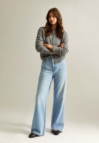 Szary sweter z dzianiny z guzikami z przodu, w połączeniu z jasnoniebieskimi spodniami o szerokich nogawkach. Czarne buty dopełniają stylizację. Neutralne tło.