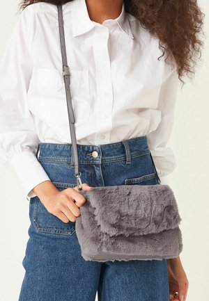 Sac en fausse fourrure grise en bandoulière avec un design à rabat, doté d'une bandoulière grise réglable et d'une texture douce, tenu contre une chemise blanche et un jean.
