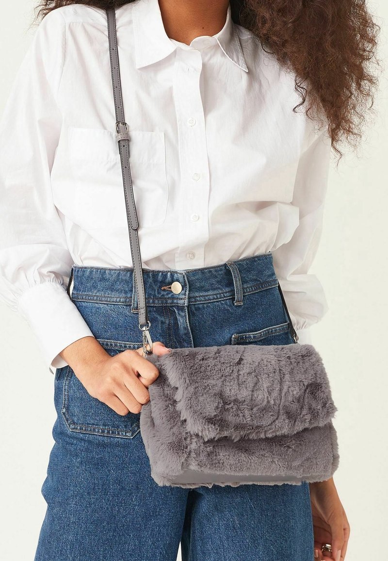 Sac en fausse fourrure grise en bandoulière avec un design à rabat, doté d'une bandoulière grise réglable et d'une texture douce, tenu contre une chemise blanche et un jean.