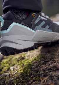 Gros plan sur une chaussure de randonnée Gore-Tex noire et grise posée sur un sol forestier moussu et irrégulier.
