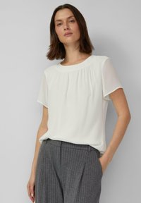 Frau trägt eine weiße Bluse mit kurzen Ärmeln und hoch taillierte graue Nadelstreifenhose, steht mit einer Hand in der Tasche vor einem schlichten Hintergrund.