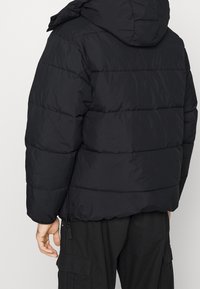 Svart vadderad jacka med huva, quiltad design och synliga sömmar. Har en exponerad dragkedja på sidan och elastiska muddar.