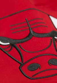 Tela roja con un destacado logo de toro bordado en negro, detallado con características faciales y acentuado por elementos blancos.