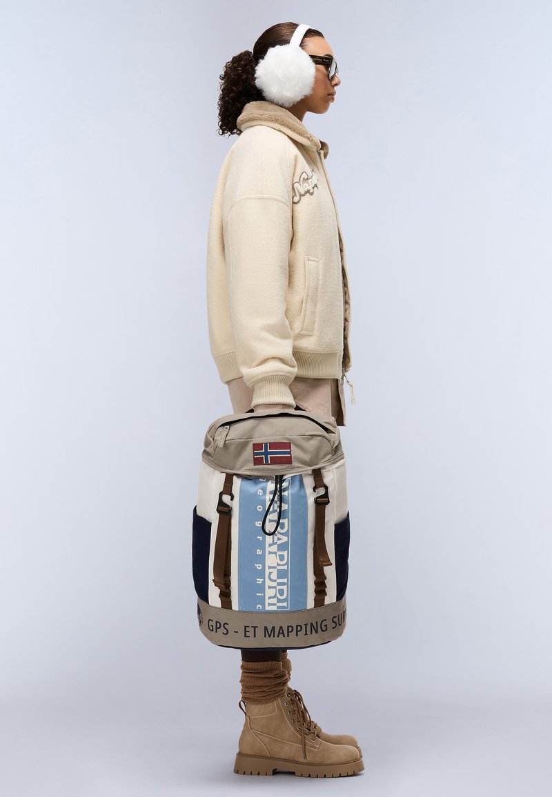 Beige Fleecejacke, brauner Pullover, blauer und beiger Rucksack mit norwegischer Flagge, dunkle Hose und beige Stiefel, mit weißen Kunstfell-Ohrenschützern.