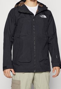 Chaqueta negra impermeable con capucha, que cuenta con un zipper frontal, dos grandes bolsillos y detalles de logotipo, fabricada con material suave y ligero.