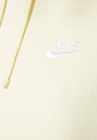 Gul hoodie i mjukt tyg, har en dragsko på huvan och en vit broderad Nike-logotyp på bröstet. Synlig strukturerad yta.