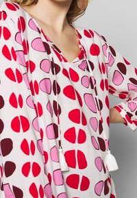 Tunique à col en V avec un fond blanc, ornée de motifs de pois abstraits en rouge, rose et bordeaux. Comprend un détail à frange au niveau du col.