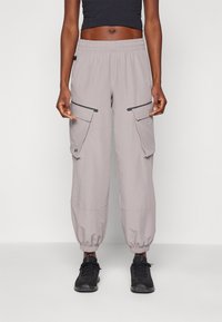 Person som bär ljusgrå cargo-joggerbyxor med elastiska anklar och svarta sneakers, stående mot en enkel ljus bakgrund.