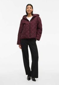 Burgundy quilted jacket with a hood, featuring a front zipper and diagonal stitching pattern. Paired with black wide-leg pants and heels.