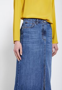 Traper suknja srednje plave boje s ravnim krojem, prednjim prorezom i detaljima od srebrne boje. Nosiva uz žutu bluzu s dugim rukavima.