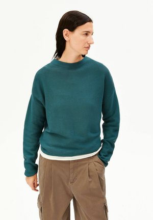 Femme aux cheveux foncés portant un pull bleu sarcelle, un sous-vêtement blanc et un pantalon marron ample, debout, une main dans la poche, regardant vers la droite.