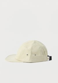 Μπεζ καπέλο πέντε πάνελ από κορδούρι με ασημένιες οπές και μαύρη ρυθμιζόμενη πόρπη σε λευκό φόντο.