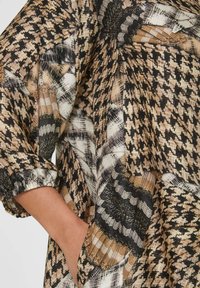 Musterbluse mit schwarzen, beigen und weißen Texturen, locker geschnitten und mit leicht gerafften Ärmeln. Enthält ein Detail mit einer Seitentasche.