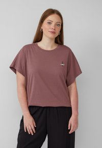 Kurzarm, lockeres T-Shirt aus mauvefarbenem Baumwollstoff mit einem kleinen gestickten Design auf der Brust. Glatte Textur, runder Ausschnitt.