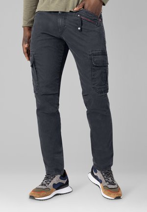 Mann trägt dunkle Cargo-Hosen und mehrfarbige Sneakers, eine Hand in der Tasche, vor einfarbigem Hintergrund.