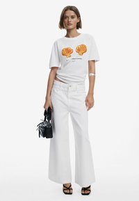 Hvid grafisk t-shirt med orange skaldtryk, parret med højtaljede brede hvide jeans og en lille sort håndtaske.