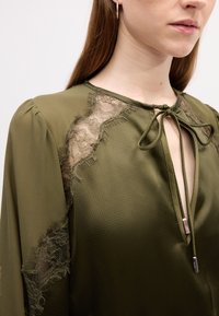 Olijfgroene blouse van gestructureerde stof met kantaccent en strikdetail bij de hals, met pofmouwen en een iets losse pasvorm.