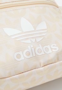 Beige tyg med ljus blommönster, med vit Adidas trefoil-logotyp och text på en dragkedjeficka.
