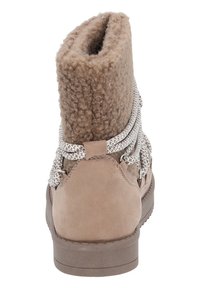 Brun mockaskinnsstövel med en texturerad, fluffig topp, prydd med vita och bruna flätade snören samt en platt gummisula. Bakifrån syns sömnadsdetaljer.