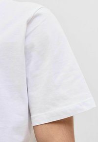Tricou din bumbac alb, cu mânecă scurtă, prim-plan al tivului și cusăturii de umăr. Textură moale, design simplu, finisaj curat cu cusătură dreaptă.