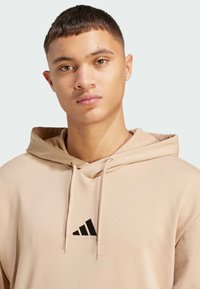 Sudadera color beige con un logo triangular negro en el pecho. Presenta una capucha con cordones y una textura suave y lisa.