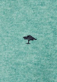 Mintgroene brei-stof heeft een klein, zwart geborduurd boomborduurwerk aan de linkerkant. De textuur lijkt zacht en casual.