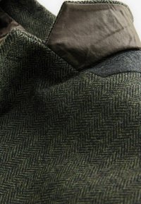 Gros plan d'une veste en laine verte à motif chevrons avec une pièce en cuir marron foncé sur le col.