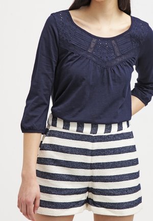 Vrouw draagt een marineblauwe blouse met kanten details en een korte broek met hoge taille en structuur, met marineblauwe en witte horizontale strepen.