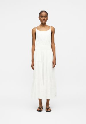 Γυναίκα που στέκεται προς τα εμπρός φοράει λευκό μακρύ φόρεμα χωρίς μανίκια με βαθμίδες και μαύρα επίπεδα σανδάλια σε λευκό φόντο.