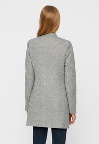 Harmaa villakangastakki, jossa on sileä rakenne, polvipituinen muotoilu ja suora leikkaus. Varustettu pitkillä hihoilla ja korkealla kauluksella.