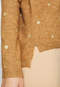 Stickad tröja i varm brun med subtila gyllene prickar. Har en ribbad mudd och en liten sidoklyfta vid fållen för extra detalj.