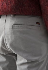 Gros plan de la poche arrière d’un pantalon gris avec une étiquette de marque BOSS noire et orange, associé à un pull rouge foncé texturé.