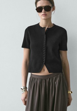 Femme portant un cardigan noir à manches courtes boutonné, une jupe plissée sombre, de grandes lunettes de soleil, et des bracelets argentés et dorés, debout devant un fond uni.