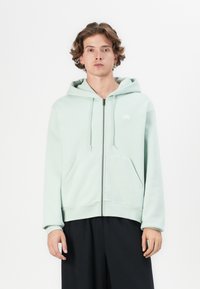 Light mint green zip-up hoodie made of soft fabric, featuring a kangaroo pocket and a logo on the left chest, with a relaxed fit.