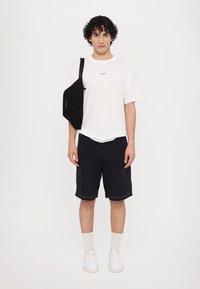 Tricou alb din bumbac cu un mic logo negru, pantaloni scurți negri, adidași albi și un rucsac negru, stând în fața unui fundal simplu.